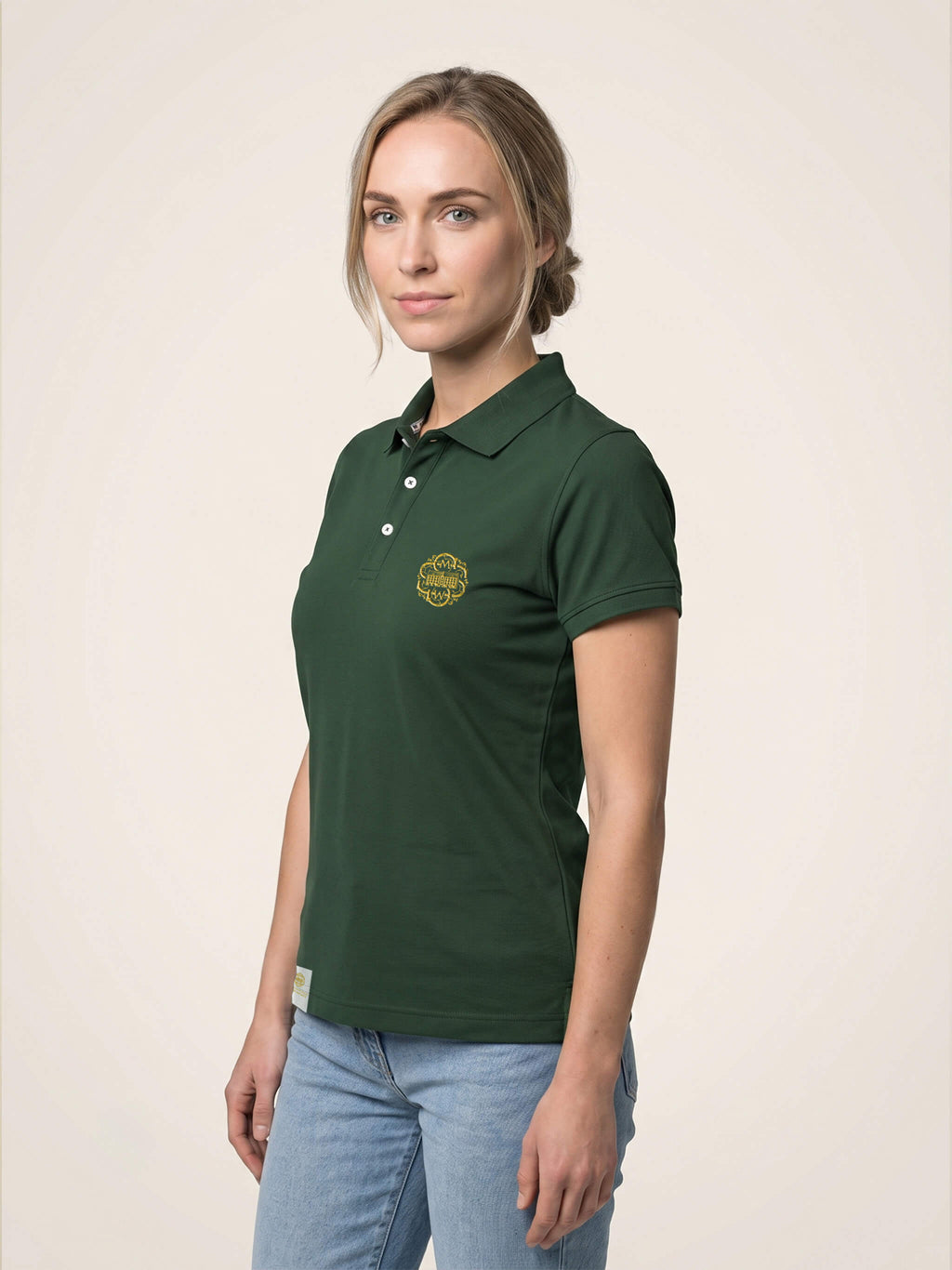 Side view of woman wearing forest green fitted polo shirt with gold SMWS logomark on the left chest.